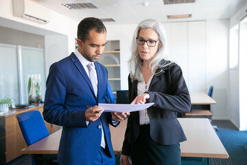 Wall Mural - Successful businessman in suit reading document for signing and female grey-haired manager in eyeglasses pointing at something in report. Partners working in office. Business and management concept