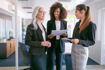 Wall Mural - Content young businesswomen discussing statistics and smiling. Three happy attractive female colleagues standing with papers and talking in conference room. Teamwork, business and management concept