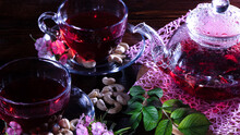 A Mug Of Red Tea And A Teapot In White Hibiscus Flowers And Green Leaves Of Medicinal Tea On A Wooden Stand.Zen Tea Ceremony. Photo Of Red Herbal Indian Healing Tea.Elegant Mugs With A Relaxing Drink