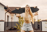 Fototapeta Londyn - Teenage girl with long blonde hair holding a skateboard on her shoulders.