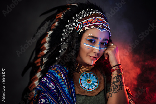 Plakat Portrait of a beautiful American Indian woman in ethnic costume and traditional make up on dark background with smoke illuminated by red light