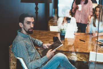 Pensive hipster guy in casual wear holding book thinking about literature during free time on break, handsome contemplative businessman puzzled on some ideas sitting at cafe interior looking away.