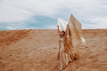 A beautiful girl in a golden with brilliant dress with wings, suit is dancing an oriental, East dance in the desert.