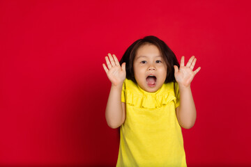 Surprising. Beautiful little girl isolated on red background. Half-lenght portrait of happy child gesturing. Cute asian girl in yellow wear. Concept of facial expression, human emotions, childhood.