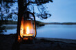 Vintage old kerosene lamp hanging on a tree. Beautiful view of glowing lantern and dark misty lake at night. Travel, Outdoor Concept