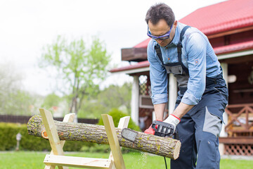 Man in overalls saws a tree on sawhorses in courtyard with a chainsaw. Cutting firewood in forest with a professional chainsaw. Hard work with a saw. Sawdust fly to sides