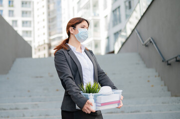 Unhappy woman in a mask is walking along the street with a box of personal stuff on the background of the stairs. A female office employee was fired. Economic crisis during epidemic covid 19.