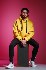 Wall Mural - A young man of 25-30 years old in a yellow sweatshirt sitting on an old music column on pink wall background and showing different emotion.