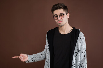 Handsome young adult man in shirt looking at camera and pointing away while standing isolated on brown background