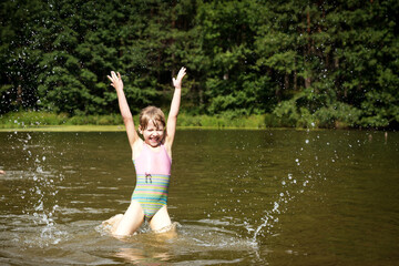 a little girl in a bathing suit jumps and splashes water in the lake in summer