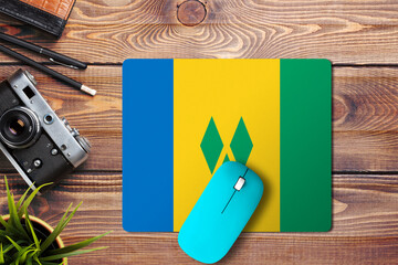 Wall Mural - Saint Vincent And The Grenadines flag on wooden background with blue wireless mouse on a mouse pad, top view. Digital media concept.