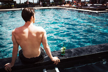 Wall Mural - Young hot man resting at swimpool. Back view of guy sitting at water alon and look forward. Enjoy his time lonely in resort spa place. Summertime period. Strong powerful man.