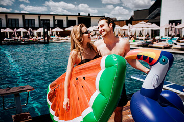 Wall Mural - Young hot couple resting at swimpool. Spend time together at water. Woman and man holding two swim ring and look at each other. Happy positive couple has rest at spa resort place.