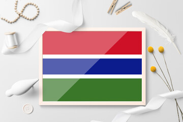 Gambia flag in wooden frame on white creative background. White theme, feather, daisy, button, ribbon objects.