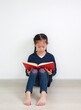 © zilvergolf - Asian little girl in school uniform reading a book and sitting against white wall in the room.