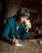 Woman makes a note on granite counter top in her kitchen as she sips a mug of tea or coffee.