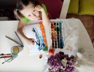 caucasian little girl child draws paints at the table top view with white cat and bouquet of flowers