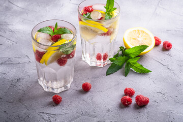 Fresh cold ice water drink with lemon, raspberry fruits and mint leaf in two faceted glass on stone concrete background, summer diet beverage, angle view selective focus