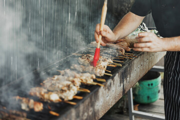 Blurred hands of man lubricates the meat with a sauce brush on long rectangular barbecue with smoke on the street outdoor. Summer barbecue cooking. Barbecue with raw vegetables. BBQ on the terrace