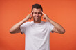 © timtimphoto - Unhappy young handsome brown haired man with short haircut keeping fingers on temples and frowning face while standing over orange background in white basic t-shirt