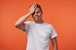 © timtimphoto - Stressed young short haired brunette guy keeping his eyes closed while frowning face and holding palm on forehead, standing isolated over orange background