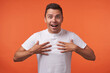 © timtimphoto - Studio photo of young pretty brunette male with short haircut looking excitedly at camera with wide smile and keeping raised hands on his chest, standing over orange background