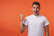 © timtimphoto - Agitated young pretty brown haired male keeping hand raised while showing aside and looking amazedly at camera with opened mouth, isolated over orange background