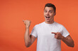 © timtimphoto - Indoor photo of young short haired brunette male looking emotionally at camera while pointing aside with raised hands, standing over orange background