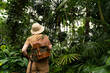 Woman botanist dressed in safari style in greenhouse, back view. Naturalist in khaki clothes, leaver gloves with backpack walks in the rainforest surrounded by palms. Jungle tourist