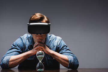 Wall Mural - Man in vr headset sitting near hourglass on table on grey background, concept of time management
