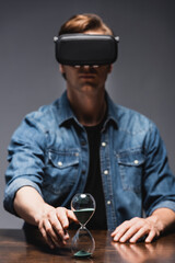 Wall Mural - Selective focus of man in virtual reality headset pulling hand to hourglass on table on grey background, concept of time management