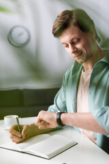 Wall Mural - Selective focus of smiling freelancer checking time while writing on notebook near cup of coffee, concept of time management