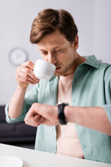 Wall Mural - Selective focus of handsome man drinking coffee and checking time on wristwatch at home, concept of time management