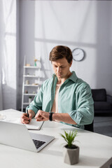 Wall Mural - Selective focus of freelancer writing on notebook and looking at wristwatch near laptop on table, concept of time management