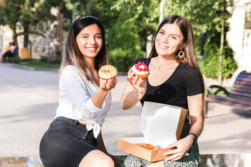 portrait of two beautiful girls friends enjoying delicious donuts with caramel and raspberry in city park. junk but tasty food for positive mood