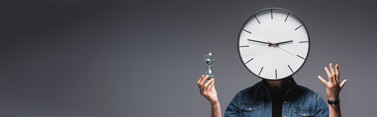 Wall Mural - Panoramic shot of man with clock on head holding hourglass and gesturing on grey background, concept of time management