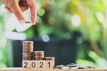 A hand putting the coins on the wooden table like a growing graph. with text 2021, On green blurry bokeh background. And copy space. Saving money and finance concept.