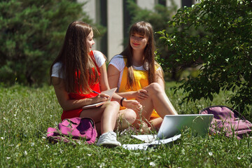 two teenage girls twins in a yellow and red school dress with purple backpacks sit outside the school in the afternoon on the lawn with books and a laptop after quarantine