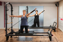 Sportswomen In Activewear Doing Exercises On Pilates Machine And Pumping Muscles With Metal Resistance Equipment