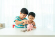 © ake1150 - Two Asian cute brother and son insert coin  in piggy bank together on the table in living room at home. Concept of bonding of sibling, friendship and saving, investment money concept