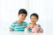 © ake1150 - Two Asian cute brother and son insert coin  in piggy bank together on the table in living room at home. Concept of bonding of sibling, friendship and saving, investment money concept