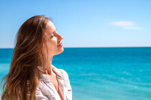 Portrait Of A Beautiful Woman With Long Hair With Closed Eyes Enjoying The Sun On Background Of A Cloudless Blue Sky And Turquoise Mediterranean Sea.