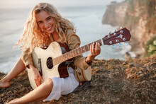 Beautiful Young Woman Playing Guitar On Beach At Sunset, Fashion Lifestyle, Look At The Camera And Smile. Happy Blond Curly Hair Womnan With Guitar Sit On Sea Cliff