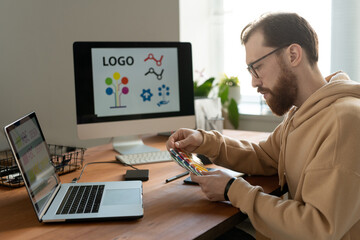 Wall Mural - Serious bearded designer in eyeglasses sitting at desk with computers and viewing color swatch while working on logo design