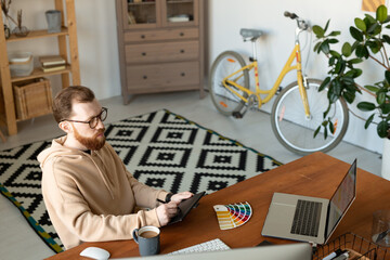 Wall Mural - Busy illustrator in hoodie sitting in home office and making sketch using drawing tablet