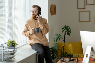 Wall Mural - Serious young bearded man in glasses sitting on window-sill and looking through blinds while communicating by phone
