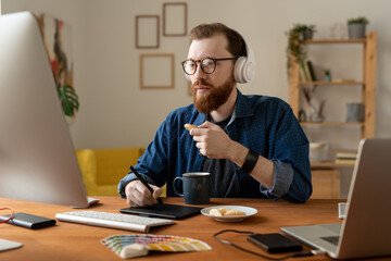 Wall Mural - Busy young bearded designer in wireless headphones sitting at table in home office and eating cookies while working with computer and digitizer