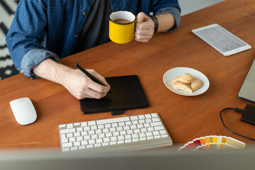 Wall Mural - Close-up of unrecognizable designer drinking coffee and using drawing tablet while working on computer graphics