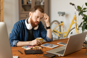 Wall Mural - Serious young bearded man focused on design concept sitting at desk and looking at sketch on laptop screen