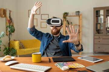 Wall Mural - Content curious bearded man sitting at desk in home office and gesturing hands while testing new app in virtual reality goggles